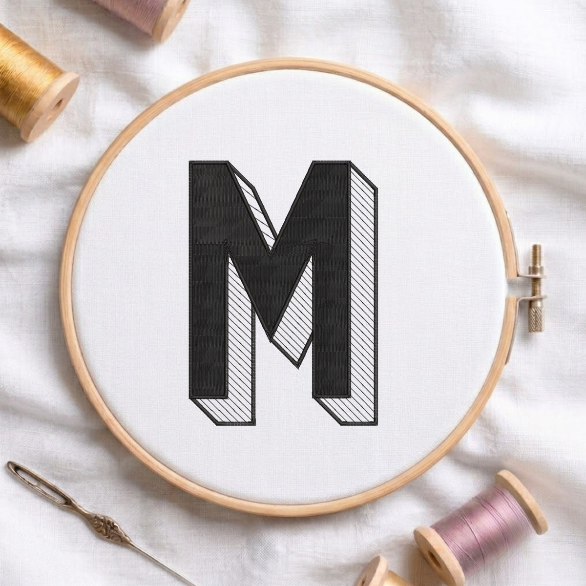 A wooden embroidery hoop holding white fabric with a large black letter M stitched inside. A needle and thread spools are arranged around it on a white fabric background.