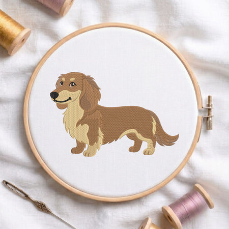A wooden embroidery hoop holds white fabric with a stitched dachshund design in warm brown and beige thread. The hoop is styled on a white fabric background with spools of thread and a needle nearby.
