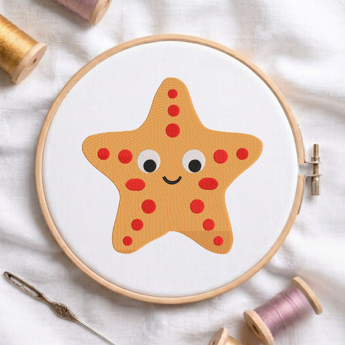 A cute cartoon yellow starfish with red spots embroidered on white fabric inside a wooden embroidery hoop, surrounded by spools of thread.