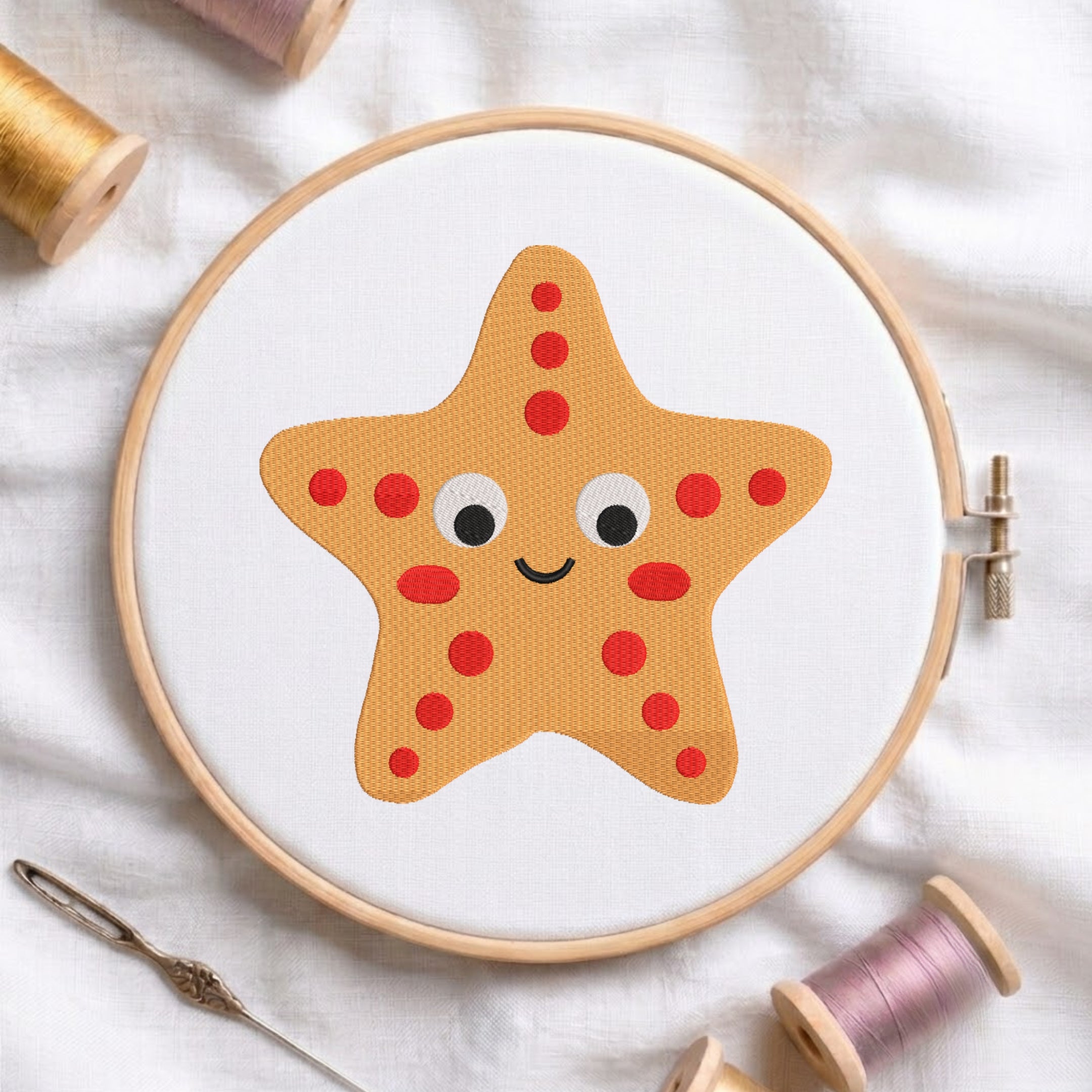 A cute cartoon yellow starfish with red spots embroidered on white fabric inside a wooden embroidery hoop, surrounded by spools of thread.