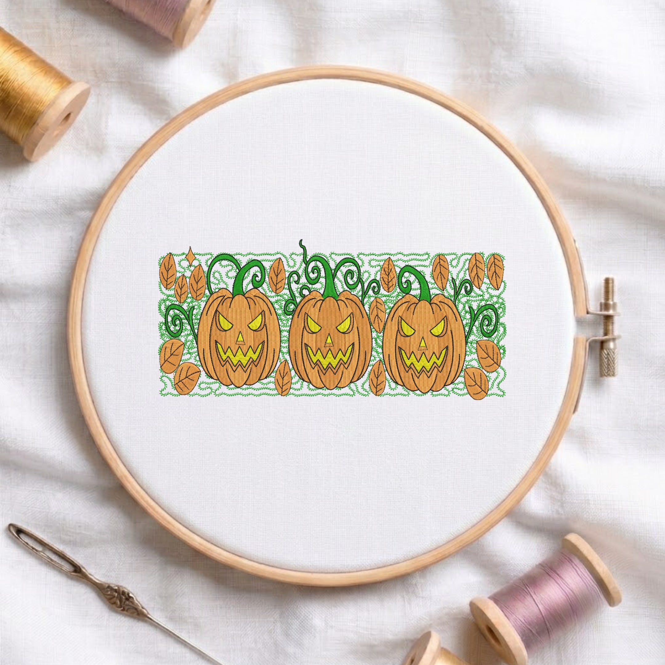 A circular embroidery hoop holding white fabric with a stitched autumn pattern of three orange jack-o'-lantern pumpkins, green vines, and scattered leaves. The hoop is styled on a white fabric background with spools of thread, a needle, and a pin around it.