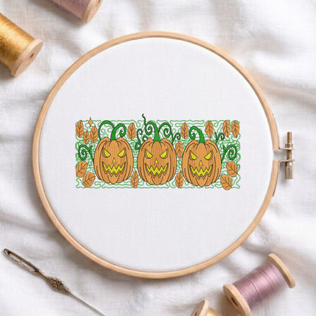A circular embroidery hoop holding white fabric with a stitched autumn pattern of three orange jack-o'-lantern pumpkins, green vines, and scattered leaves. The hoop is styled on a white fabric background with spools of thread, a needle, and a pin around it.