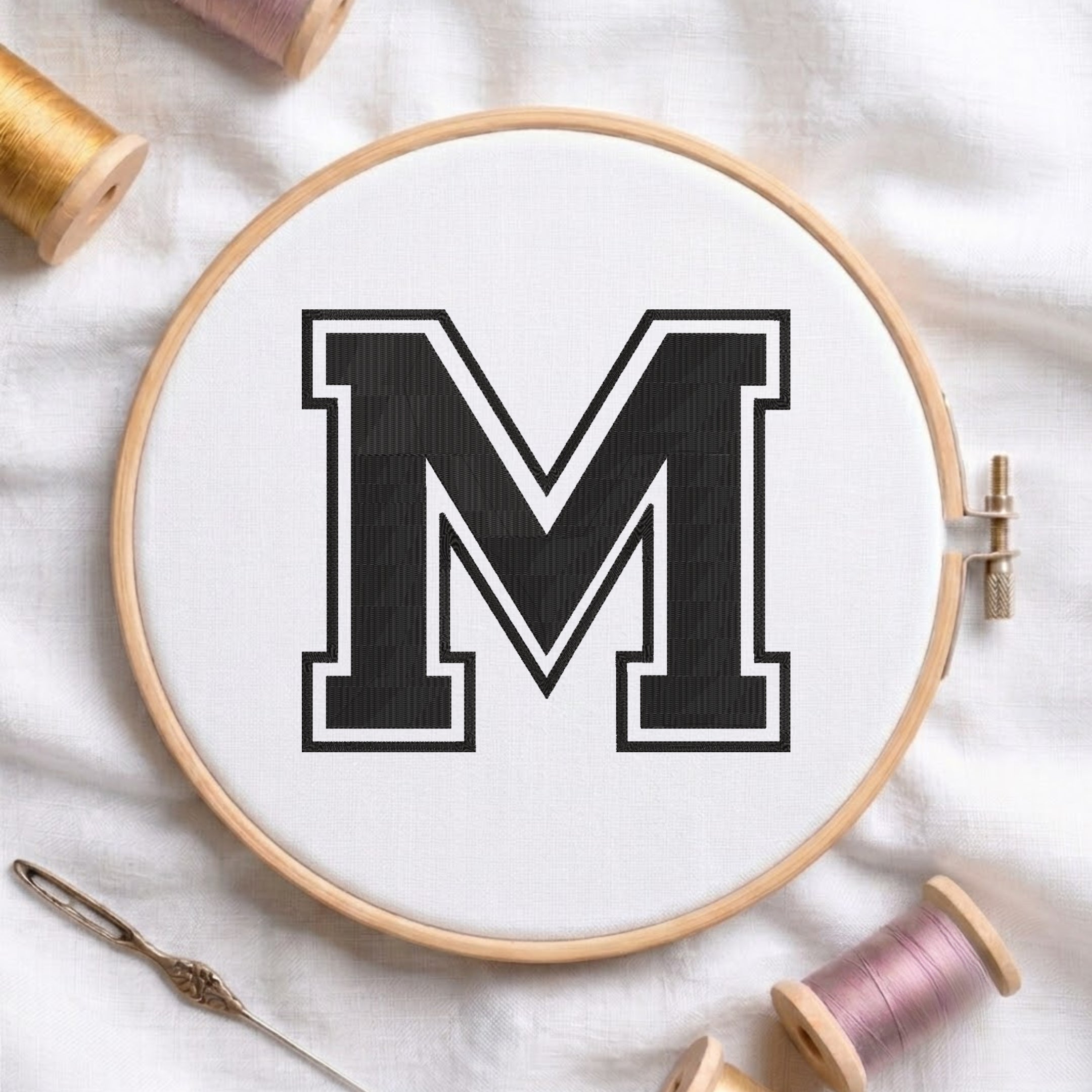 A wooden embroidery hoop holding white fabric with a large black letter M outlined in white, styled on a white fabric background with spools of thread and a needle.