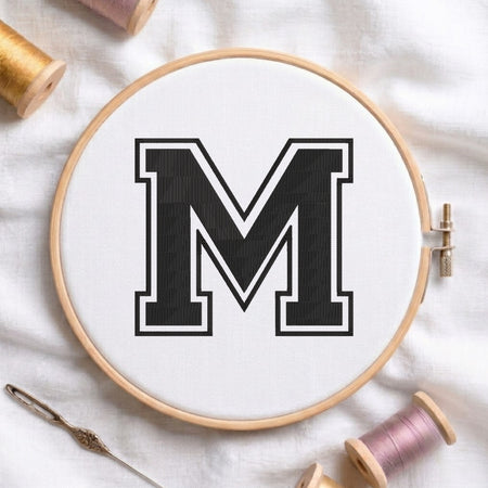 A wooden embroidery hoop holding white fabric with a large black letter M outlined in white, styled on a white fabric background with spools of thread and a needle.