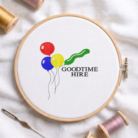 Colorful balloons and text machine embroidery design displayed in a wooden hoop on white fabric with thread spools and a needle