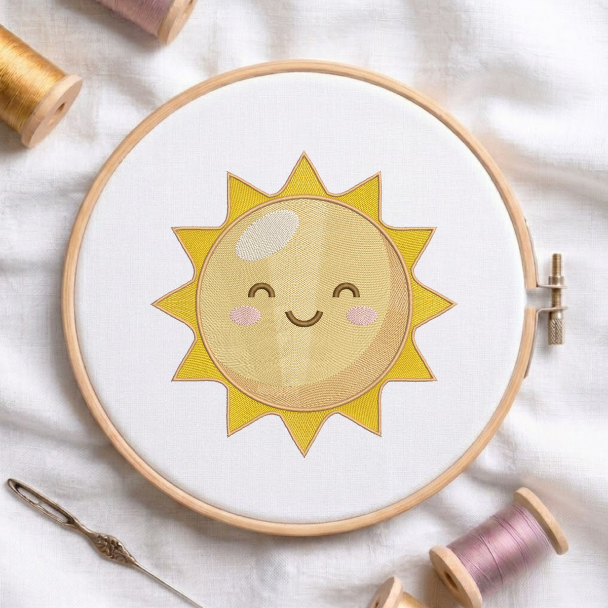 A wooden embroidery hoop holding white fabric with a smiling yellow sun applique in the center. A needle and thread spools are arranged around it on a soft white fabric background.