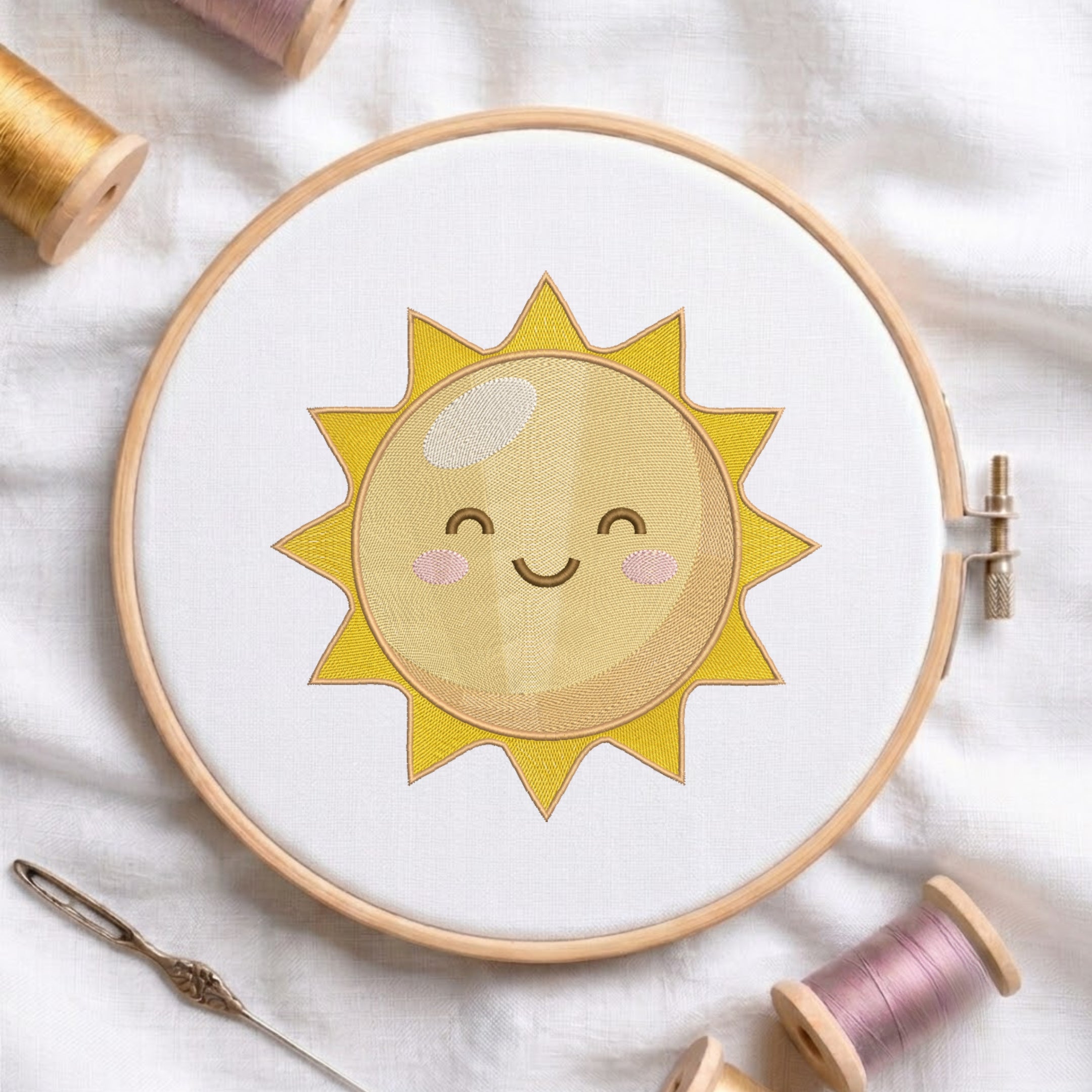 A wooden embroidery hoop holding white fabric with a smiling yellow sun applique in the center. A needle and thread spools are arranged around it on a soft white fabric background.