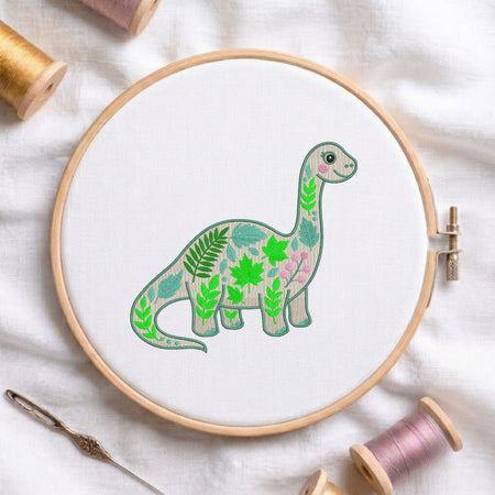 A circular embroidery hoop holding white fabric with a stylized green dinosaur design filled with leaf patterns, surrounded by spools of thread and a needle on a white fabric background.