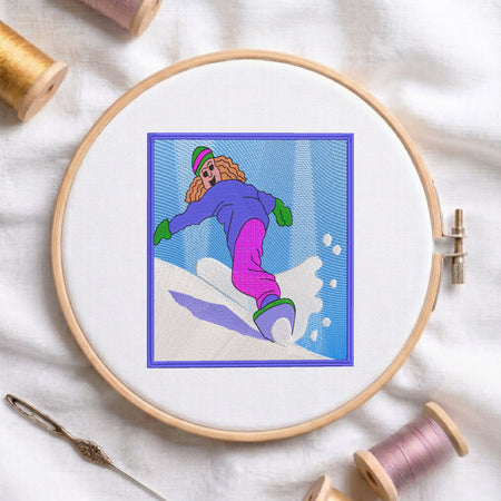 A circular embroidery hoop containing a colorful stitched illustration of a person snowboarding, surrounded by spools of thread and a needle on a white fabric background.