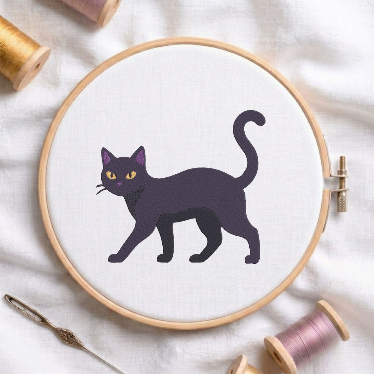 A circular embroidery hoop holding white fabric with a stylized black cat design outlined in dark purple, surrounded by spools of thread and a needle on a white fabric background.