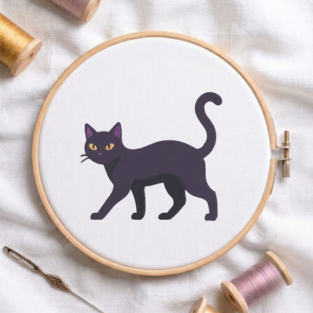 A circular embroidery hoop holding white fabric with a stylized black cat design outlined in dark purple, surrounded by spools of thread and a needle on a white fabric background.