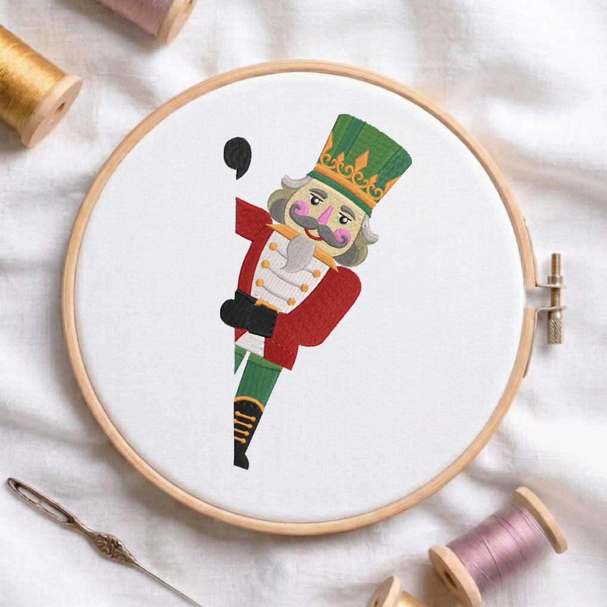 A white fabric stretched in a wooden embroidery hoop, featuring an embroidered peeking Christmas nutcracker soldier wearing a red jacket, green hat, and decorative gold trim.