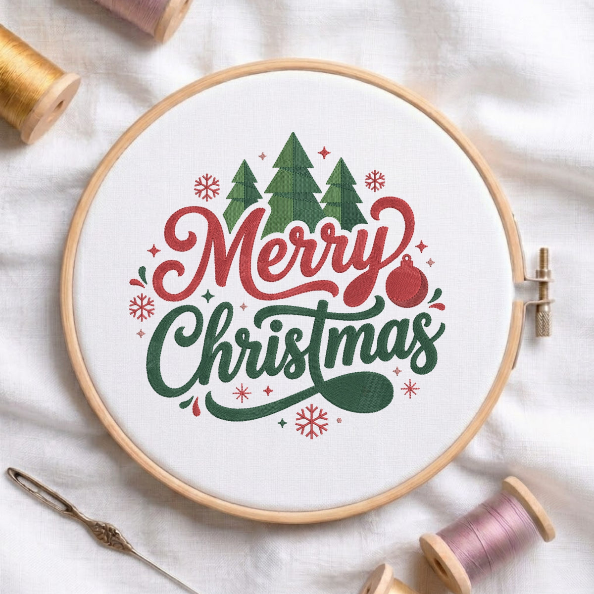 A circular embroidery hoop on white fabric, featuring a festive 'Merry Christmas' design with green trees, red lettering, and a red ornament. Spools of thread and a needle are arranged around it.