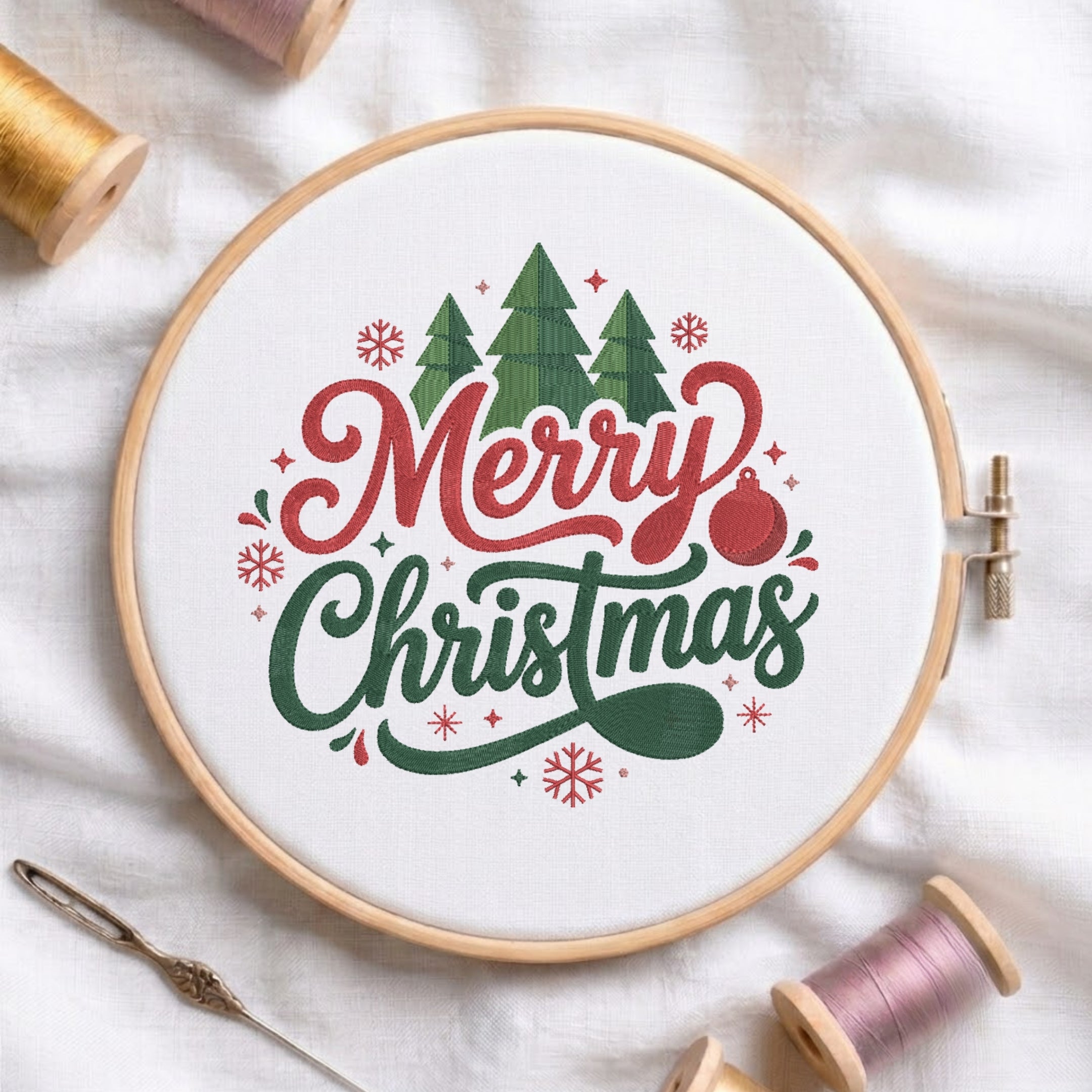 A circular embroidery hoop on white fabric, featuring a festive 'Merry Christmas' design with green trees, red lettering, and a red ornament. Spools of thread and a needle are arranged around it.