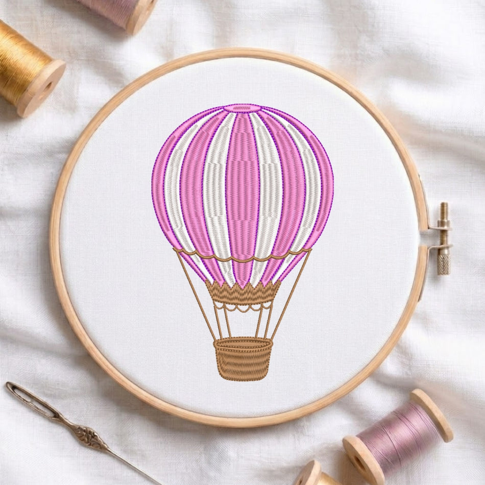 The hot air balloon embroidery design is displayed inside a wooden embroidery hoop, alongside embroidery thread spools and a needle.