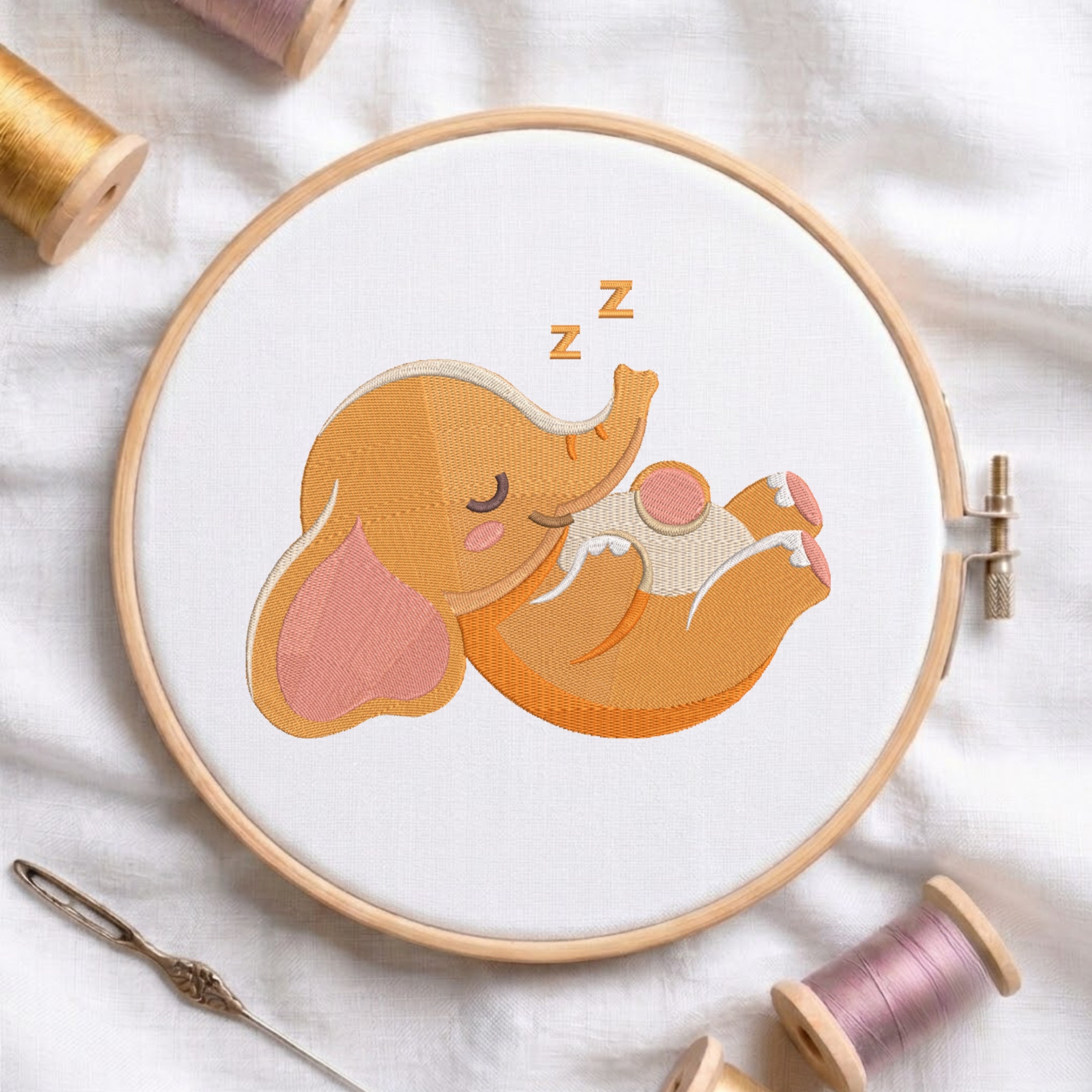 A wooden embroidery hoop holds white fabric with a stitched sleeping elephant design in orange, peach, pink, and cream tones, with small 'z' marks above its head. The hoop is styled on a white fabric surface with spools of thread and a needle around it.