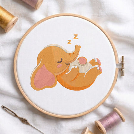 A wooden embroidery hoop holds white fabric with a stitched sleeping elephant design in orange, peach, pink, and cream tones, with small 'z' marks above its head. The hoop is styled on a white fabric surface with spools of thread and a needle around it.