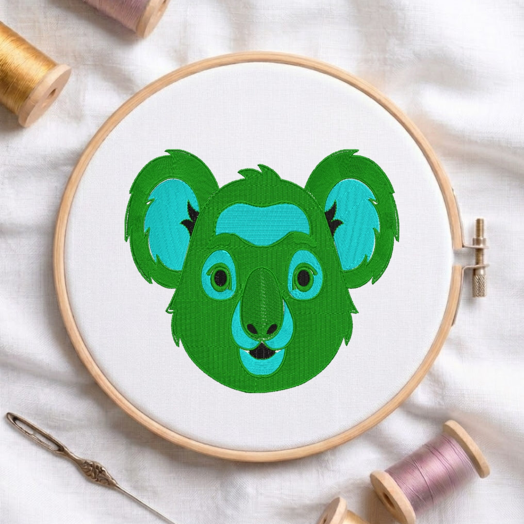 A circular embroidery hoop holding white fabric with a stylized koala face design in green and turquoise. The hoop is photographed on a white fabric surface with spools of thread and a needle around it.
