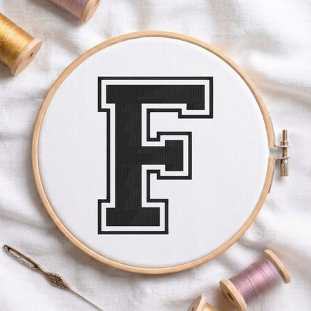 A wooden embroidery hoop holding white thread with a large black letter F outlined in white, styled on a white fabric background with spools of thread and a needle.