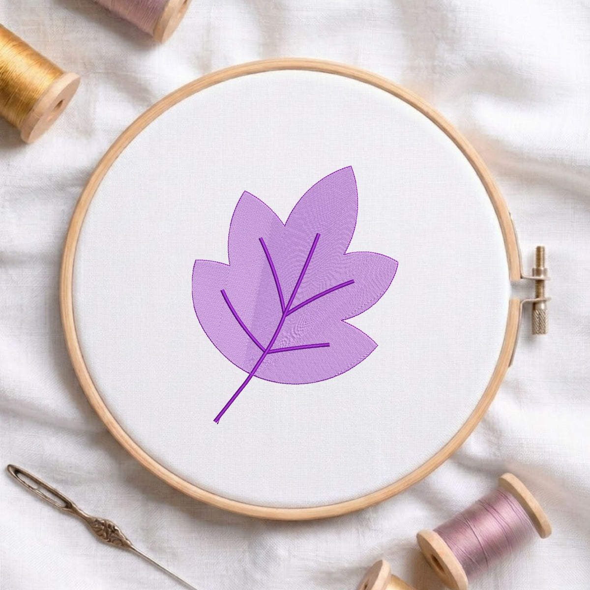 A circular embroidery hoop holding white fabric with a stitched purple leaf design, surrounded by spools of thread and a needle on a white fabric background.