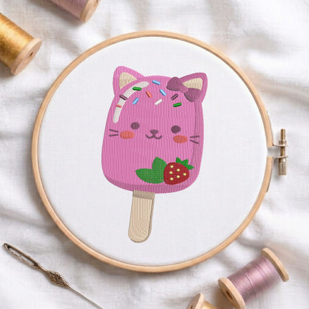 A pink cat-shaped popsicle embroidery design displayed inside a wooden embroidery hoop on white fabric, surrounded by spools of thread and a needle.