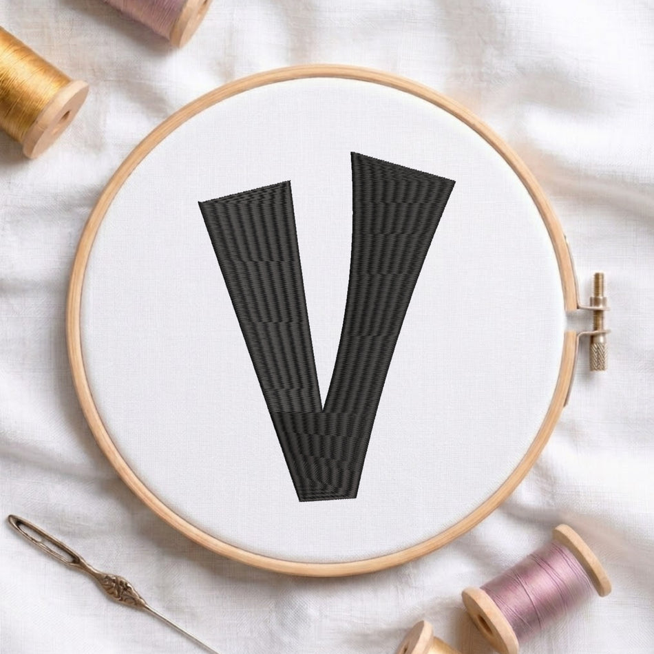 A wooden embroidery hoop holds white fabric with a large black letter V stitched in the center. Spools of thread and a needle are arranged around the hoop on a white fabric background.