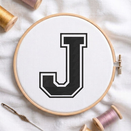 A wooden embroidery hoop holding white fabric with a large black letter J outlined in white, styled on a white fabric background with spools of thread and a needle.