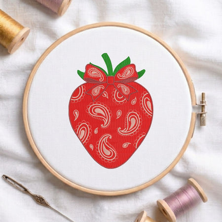 A circular embroidery hoop holding white fabric with a red strawberry design featuring white paisley patterns and green leaves, surrounded by spools of thread and a needle on a white fabric background.