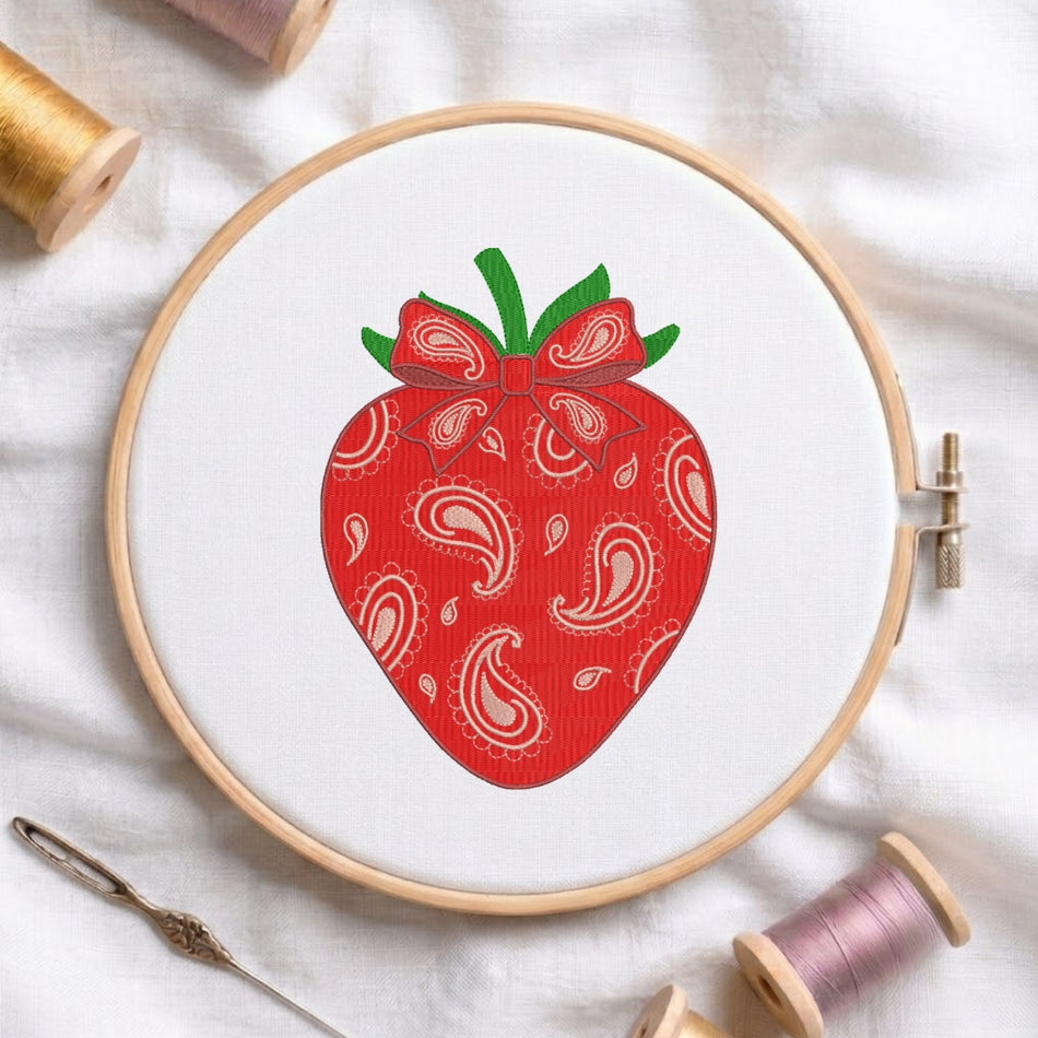 A circular embroidery hoop holding white fabric with a red strawberry design featuring white paisley patterns and green leaves, surrounded by spools of thread and a needle on a white fabric background.