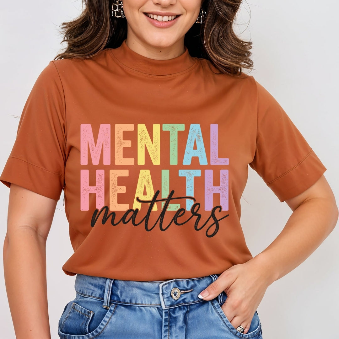 A woman wearing a rust-orange mock-neck t-shirt with the words 'MENTAL HEALTH matters' printed on the front in pastel rainbow letters and black script, paired with blue jeans.