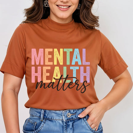 A woman wearing a rust-orange mock-neck t-shirt with the words 'MENTAL HEALTH matters' printed on the front in pastel rainbow letters and black script, paired with blue jeans.
