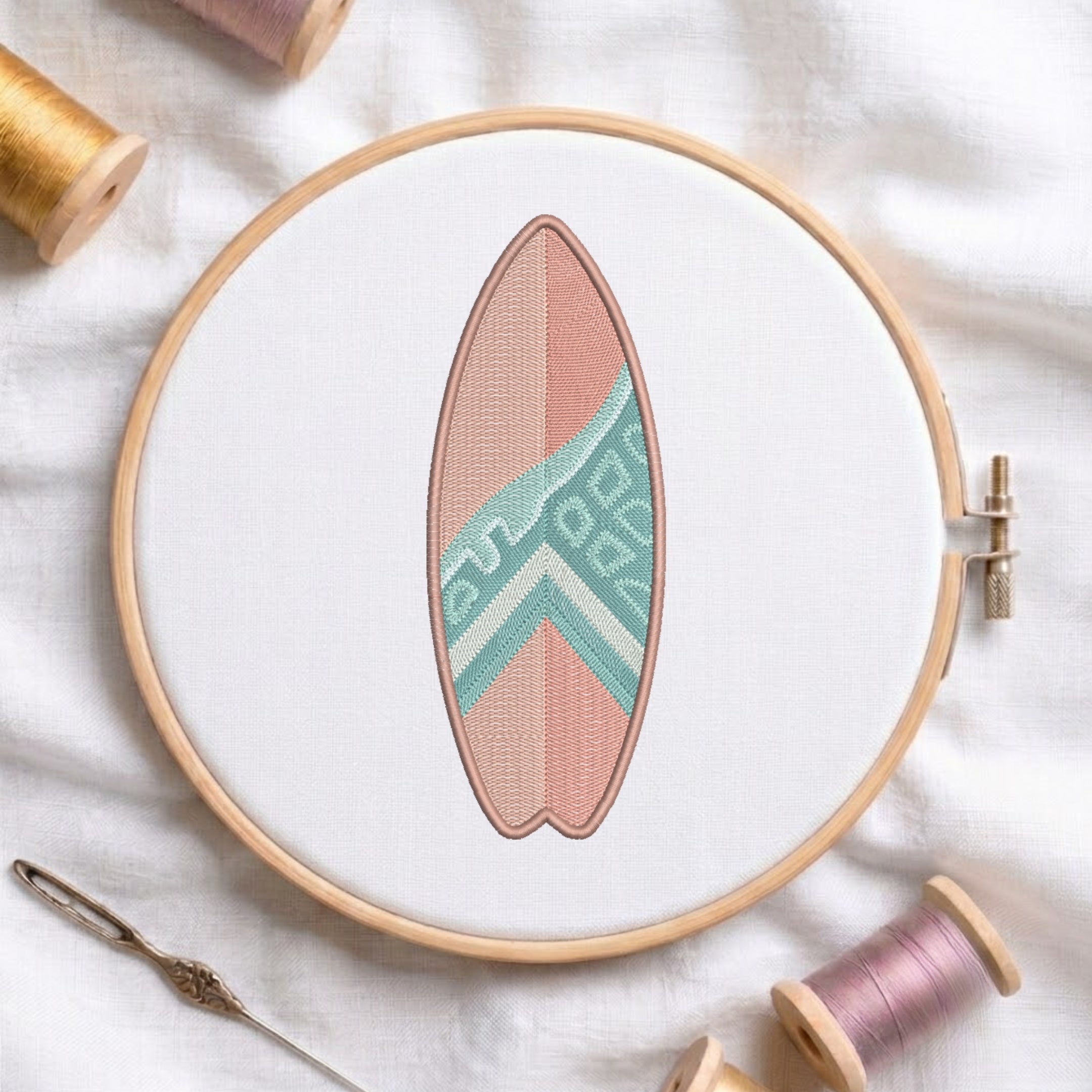 A circular embroidery hoop holds a surfboard-shaped patch or applique with a pink body and a mint green geometric chevron pattern. The hoop is styled on a white fabric background with spools of thread and a needle nearby.