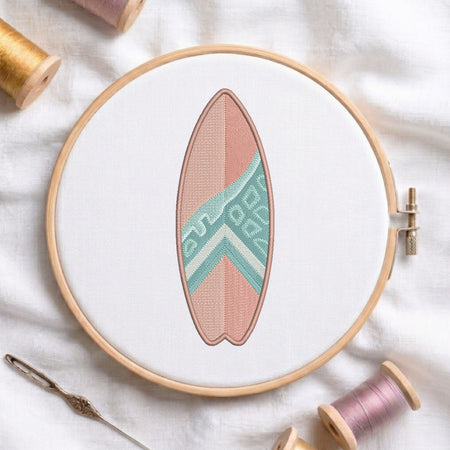A circular embroidery hoop holds a surfboard-shaped patch or applique with a pink body and a mint green geometric chevron pattern. The hoop is styled on a white fabric background with spools of thread and a needle nearby.