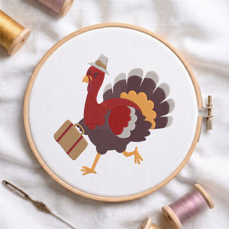 A circular embroidery hoop holds white fabric with a colorful turkey design wearing a hat and carrying a suitcase. The hoop is styled on a white fabric background with spools of thread, a needle, and wooden ends visible around it.