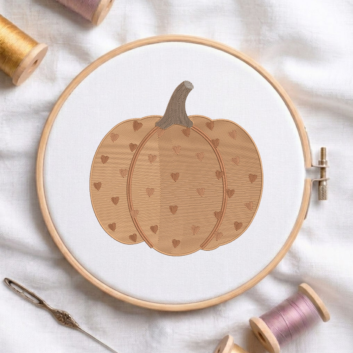 A circular embroidery hoop holds white fabric with a stitched pumpkin design in warm tan and brown tones, surrounded by spools of thread and a needle on a soft white fabric background.