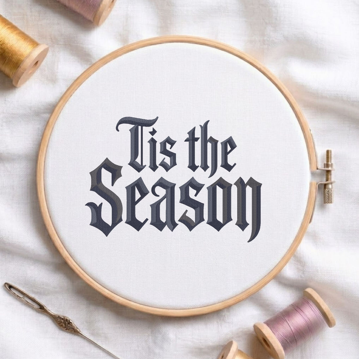 A round embroidery hoop holding white fabric with the phrase 'This is the Season' stitched in dark gray gothic lettering, surrounded by spools of thread and a needle on a white fabric background.