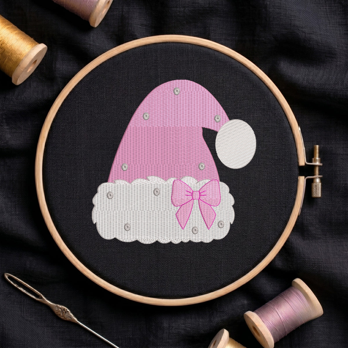 A pink and white embroidered Santa hat design stitched in a wooden embroidery hoop, surrounded by spools of thread and a needle on a dark fabric background.
