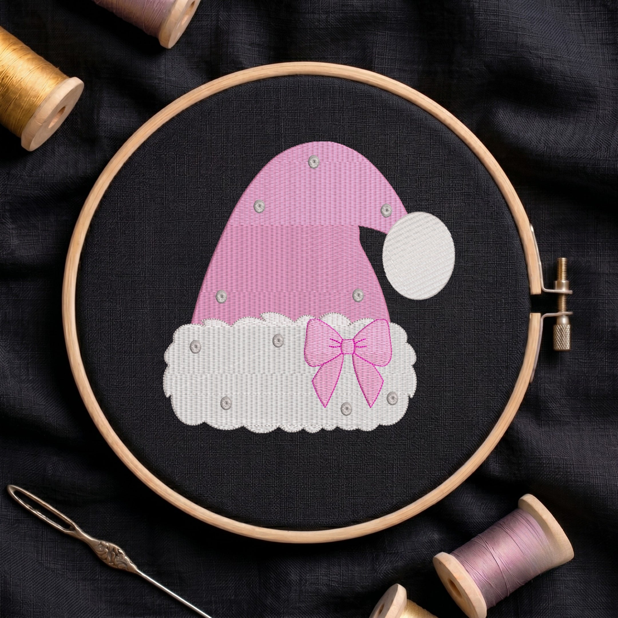 A pink and white embroidered Santa hat design stitched in a wooden embroidery hoop, surrounded by spools of thread and a needle on a dark fabric background.