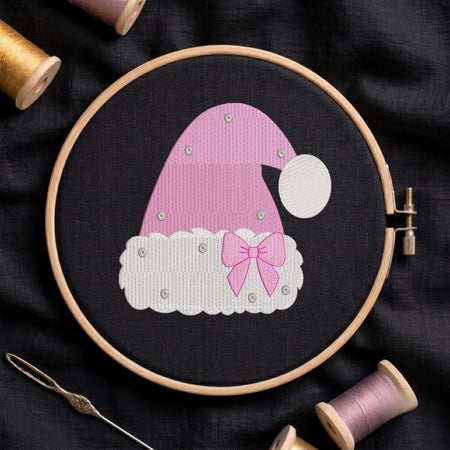 A pink and white embroidered Santa hat design stitched in a wooden embroidery hoop, surrounded by spools of thread and a needle on a dark fabric background.