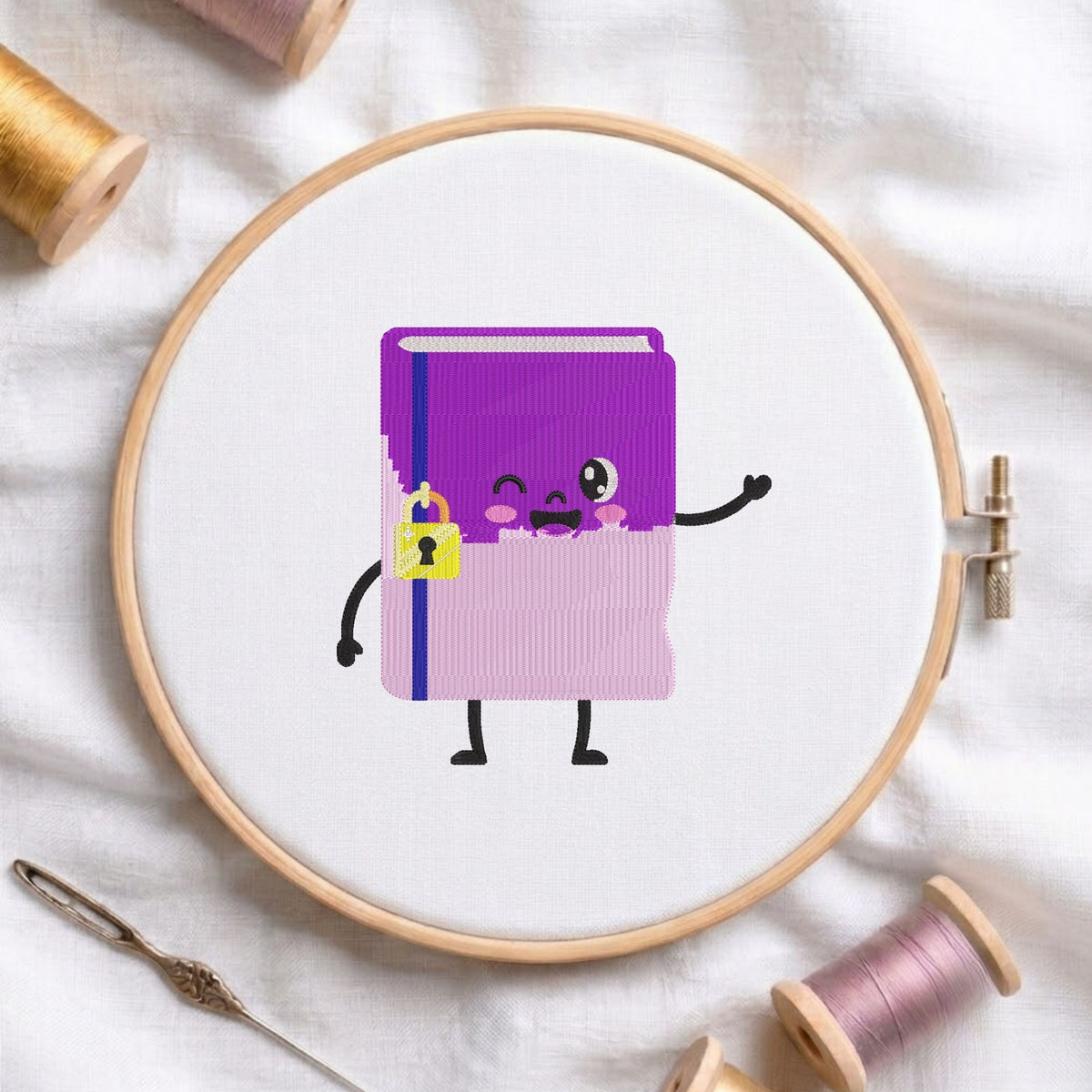 Cute purple and pink kawaii notebook character with a yellow padlock and a friendly waving hand machine embroidery design, displayed inside a round wooden hoop on white fabric with thread spools and a needle.