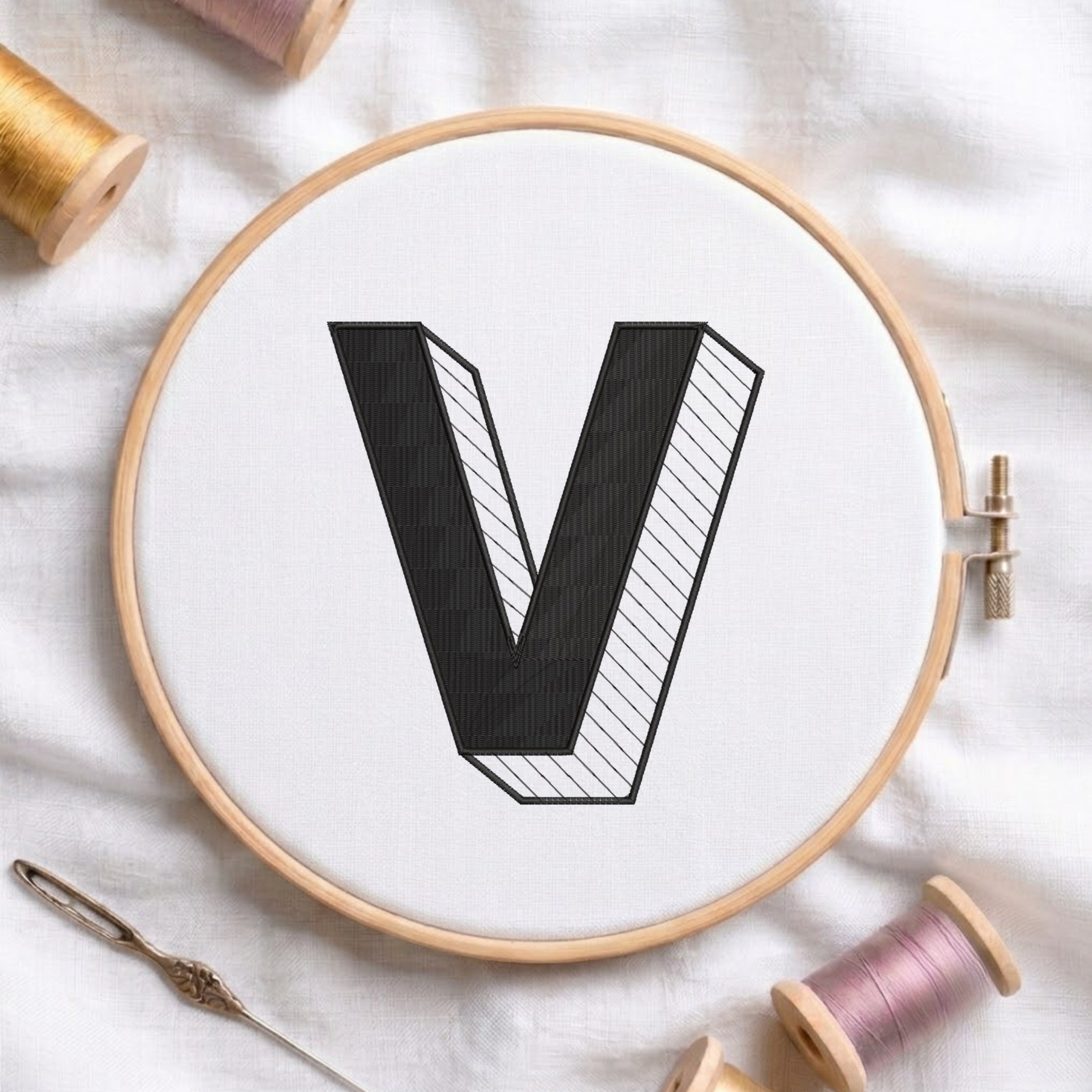 A wooden embroidery hoop holding white fabric with a large black letter V stitched in the center. A metal needle lies at the lower left, and spools of thread in gold, pink, and white are arranged around the hoop on a white fabric background.