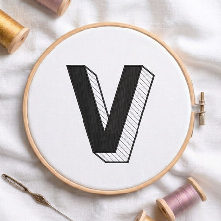 A wooden embroidery hoop holding white fabric with a large black letter V stitched in the center. A metal needle lies at the lower left, and spools of thread in gold, pink, and white are arranged around the hoop on a white fabric background.