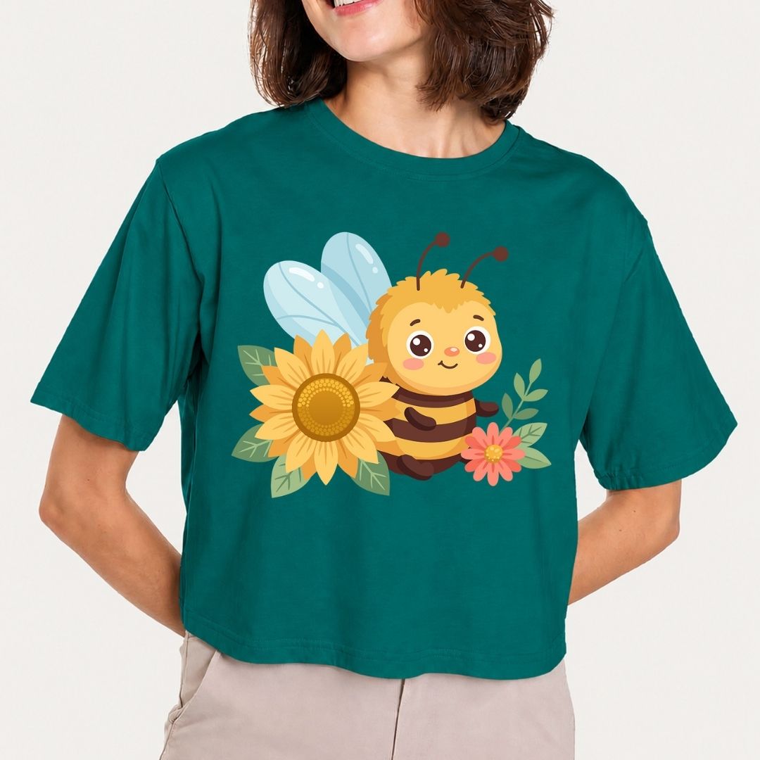 A woman wearing a dark teal cropped t-shirt with a cute cartoon bee, sunflower, and pink flower graphic on the front, paired with light beige pants against a light gray background.