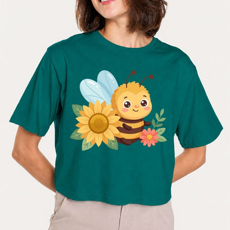 A woman wearing a dark teal cropped t-shirt with a cute cartoon bee, sunflower, and pink flower graphic on the front, paired with light beige pants against a light gray background.