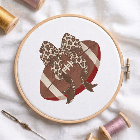 A circular embroidery hoop on white fabric, featuring a stitched design of a brown football with red and cream stripes, a leopard-print bow, and ribbon tails. Spools of thread and a needle are arranged around it on a white fabric background.