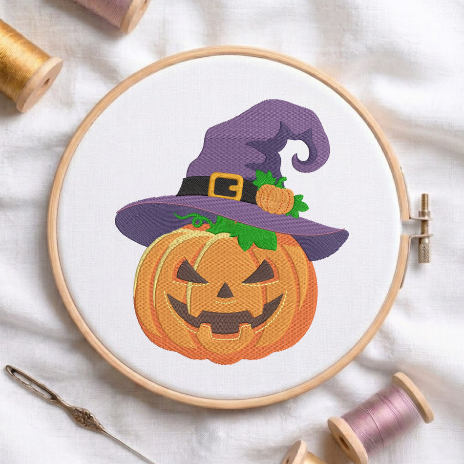 A circular embroidery hoop holding a white fabric panel with a stitched design of a smiling orange jack-o'-lantern pumpkin wearing a purple witch hat with a black band and gold buckle, surrounded by spools of thread on a white fabric background.