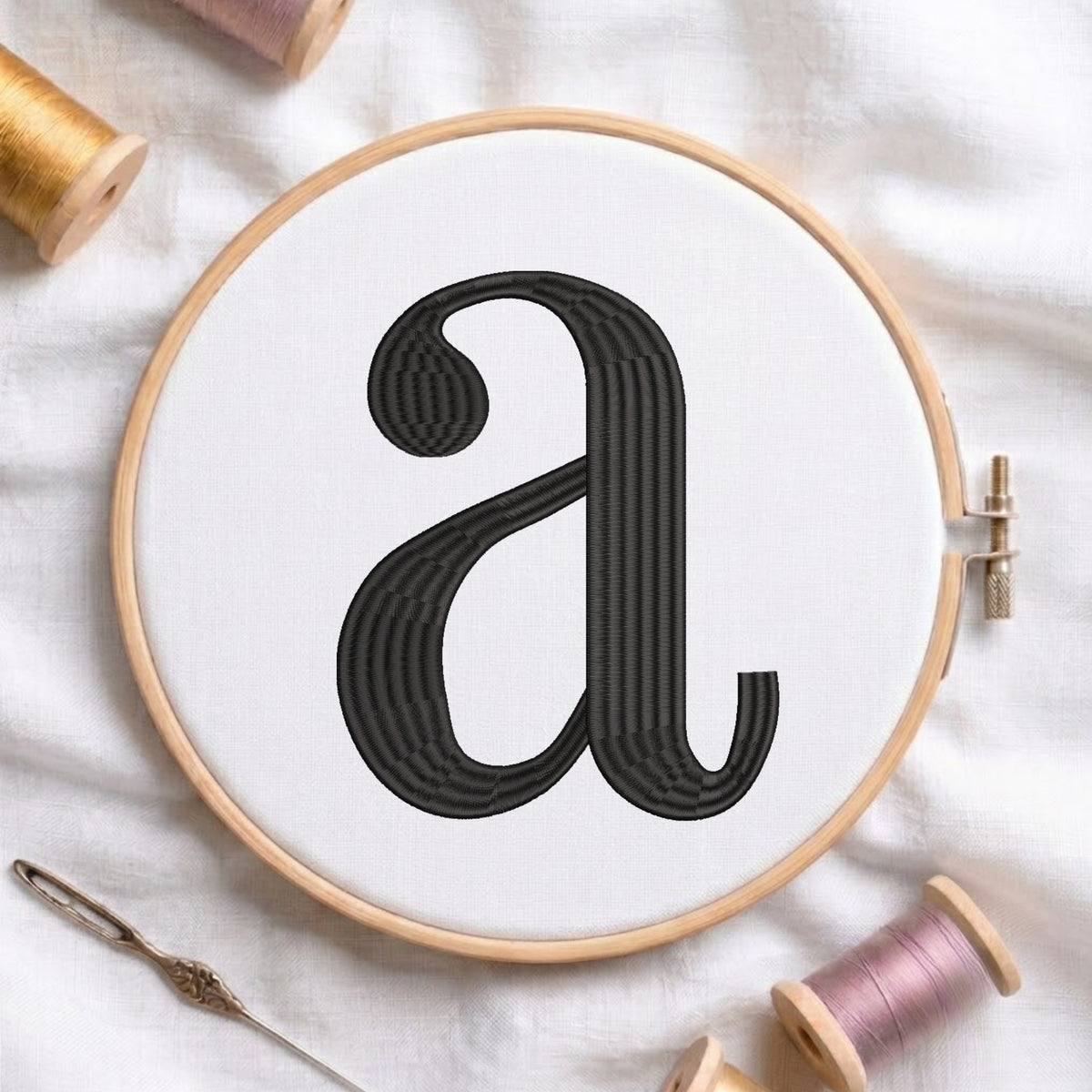 A wooden embroidery hoop holding white fabric with a large black embroidered lowercase letter 'a', styled on a white cloth with spools of thread and a needle around it.