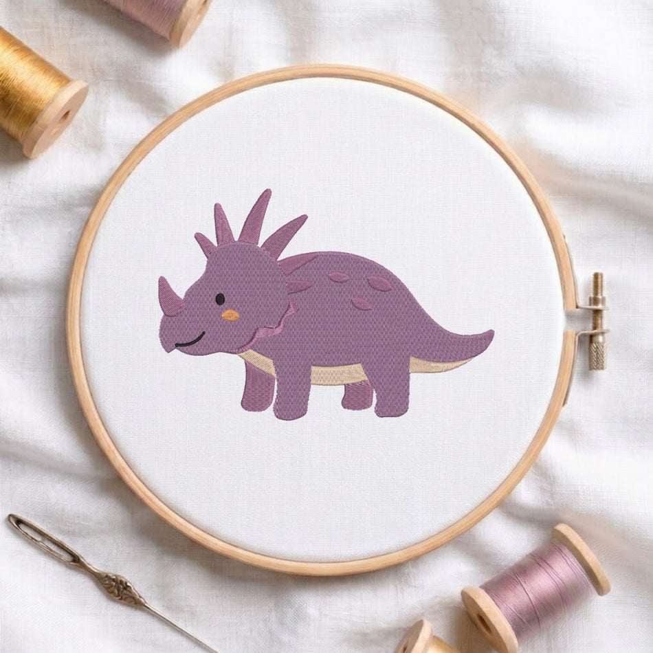 An embroidery hoop on a white cloth displays a finished embroidery design of a cute purple triceratops dinosaur with prominent head spikes and back spots, flanked by spools of thread and a needle.