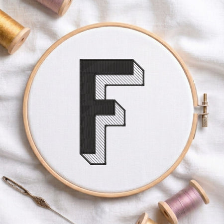 A wooden embroidery hoop holding white fabric with a large black letter F stitched in a geometric pattern. The hoop is styled on a white fabric background with spools of thread and a needle.