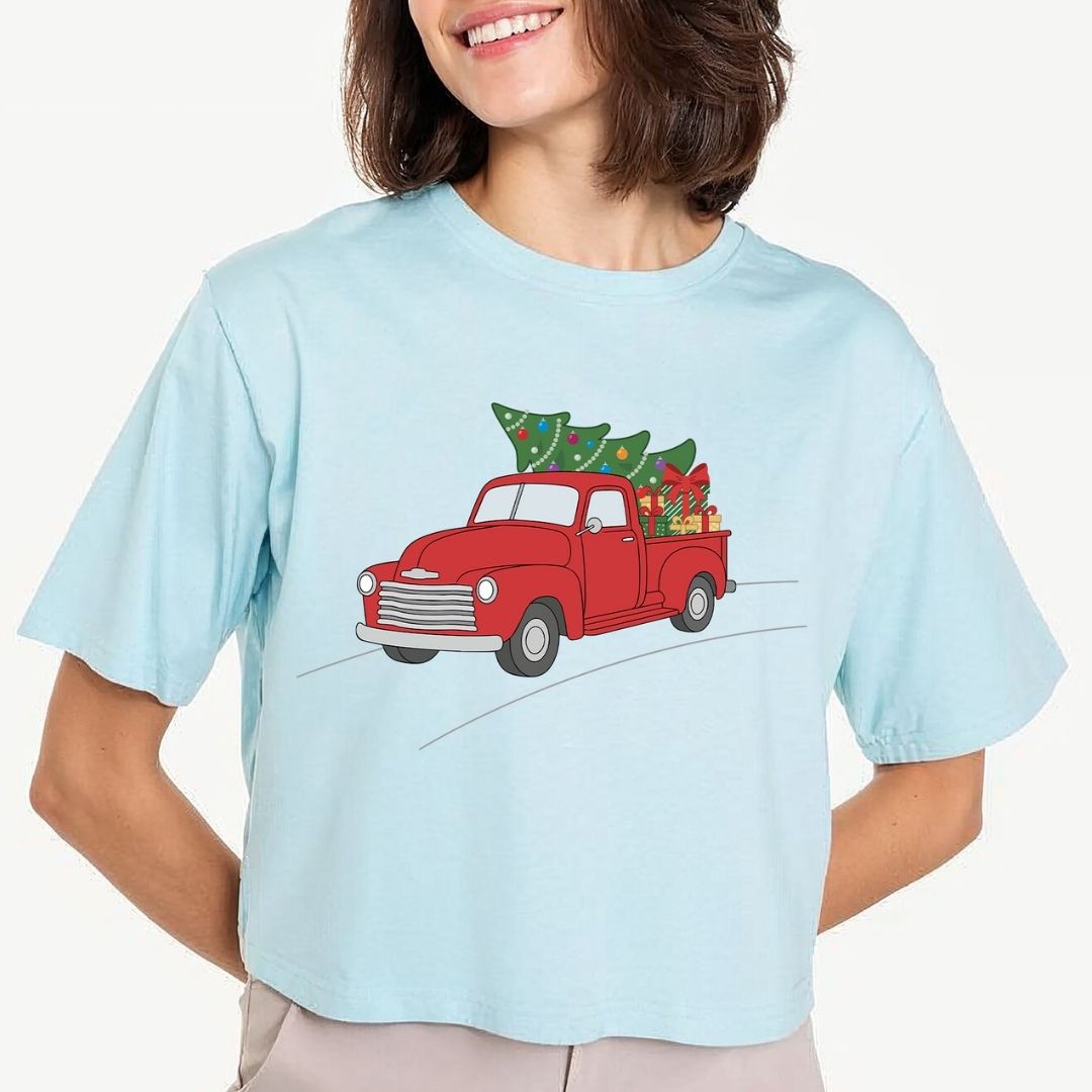 A woman wearing a light blue short-sleeve t-shirt with a graphic of a red vintage pickup truck carrying a green Christmas tree.